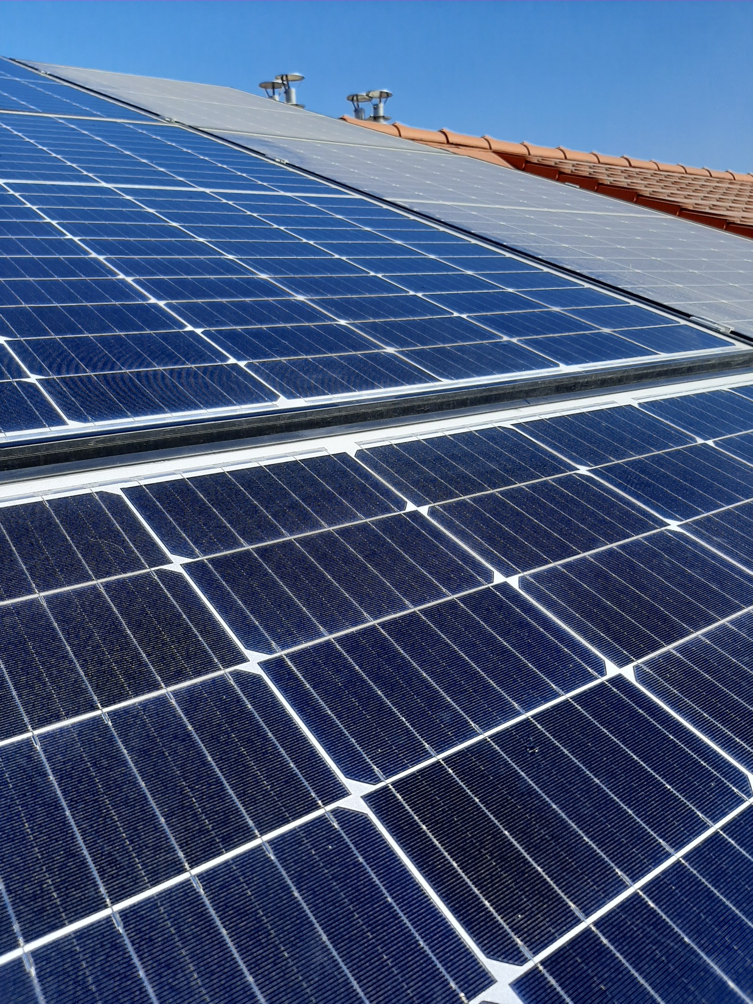 Błękitne panele słoneczne na dachu z czerwonej dachówki, widoczne kominy wentylacyjne w Mosinie, jasne niebo.