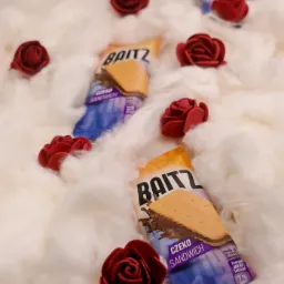Dwa batoniki czekoladowe typu sandwich, marki Baitz, ułożone wśród białych, puszystych włókien imitujących chmury, ozdobione małymi, czerwonymi różyczkami.