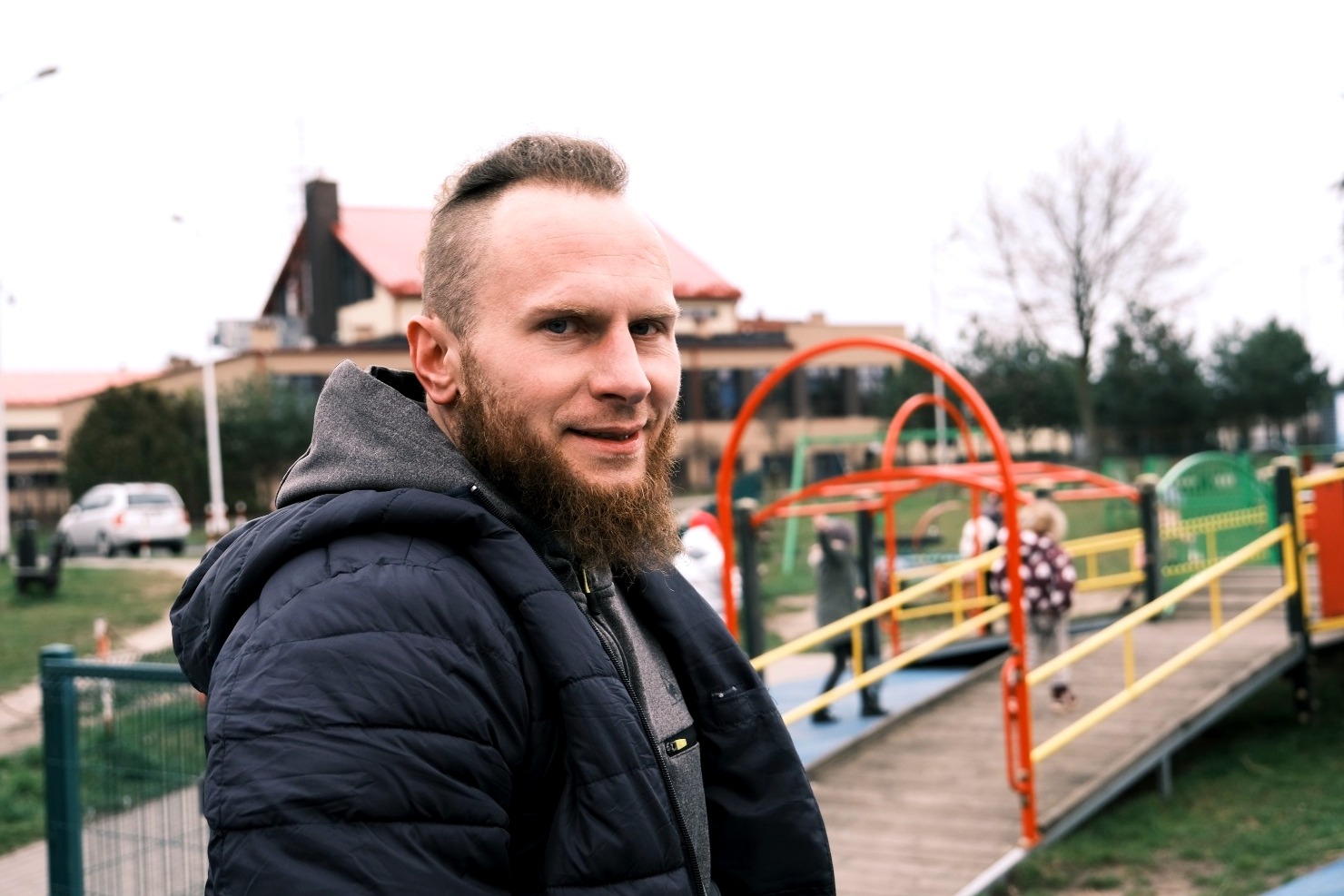 Portret mężczyzny z brodą na tle placu zabaw z dziećmi, w tle budynek z czerwoną dachówką.