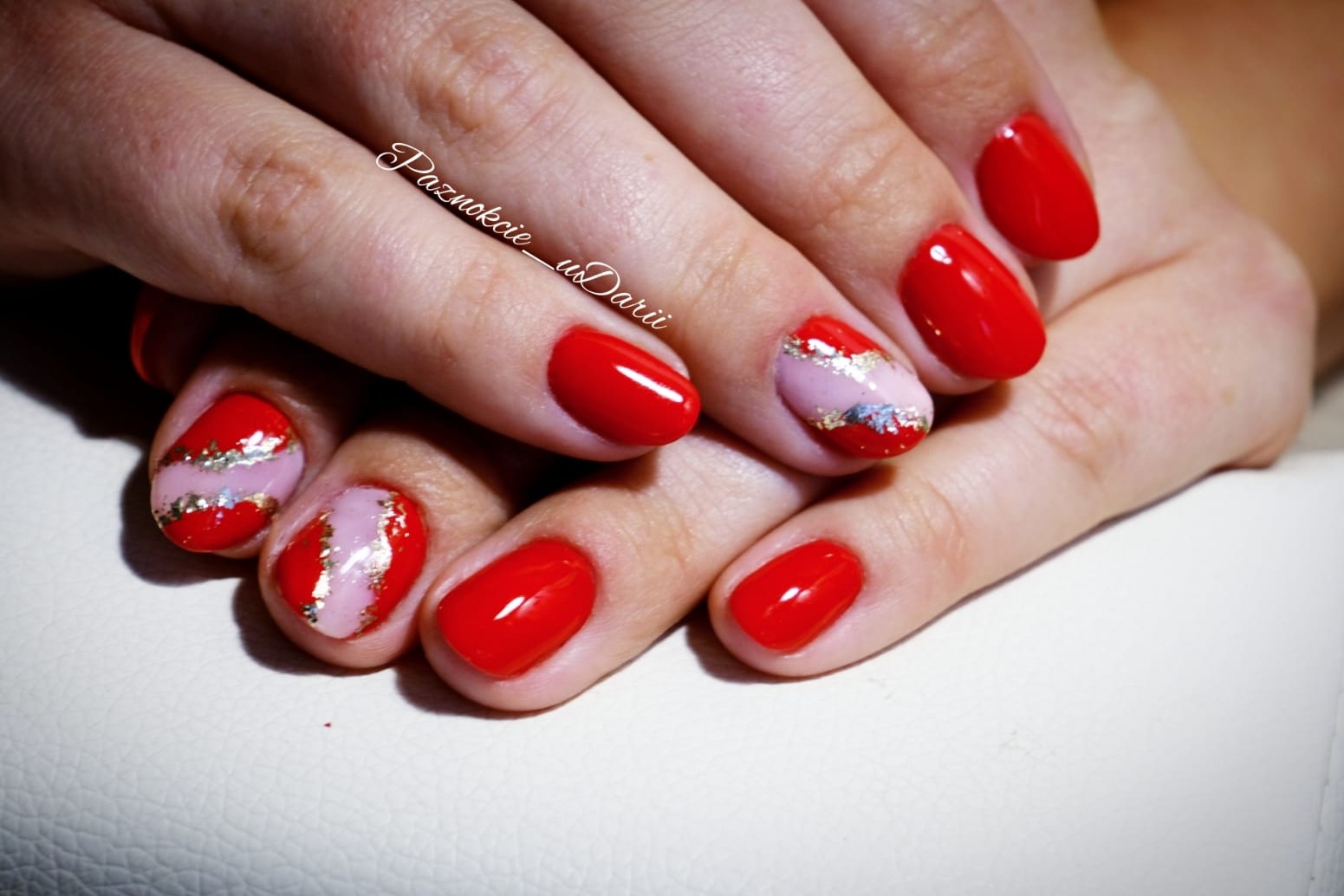 Dłonie z czerwonym manicure hybrydowym, dwa paznokcie z jasnoróżowym paskiem i złotymi płatkami.