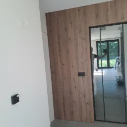 Panel ścienny z drewnopodobną okleiną i czarnymi włącznikami światła, przylegający do szklanych drzwi z czarną ramą, widok na wnętrze domu w trakcie wykańczania.