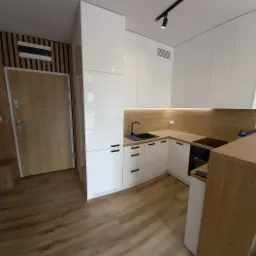 Nowoczesna, jasna kuchnia z białymi szafkami i drewnianym blatem, czarnym zlewozmywakiem i minimalistyczną armaturą, oświetlona punktowymi lampami sufitowymi i listwą LED pod szafkami wiszącymi.