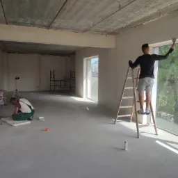 Dwóch mężczyzn pracuje w surowym wnętrzu. Jeden stoi na drabinie, wygładzając ścianę szpachelką przy oknie, drugi przygotowuje coś na podłodze. Widoczne worki z zaprawą i surowy betonowy sufit.
