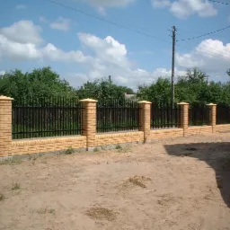 Żółta ceglana podmurówka z czarnym metalowym ogrodzeniem i filarami zwieńczonymi daszkami, widok z poziomu gruntu na tle błękitnego nieba z obłokami i zielenią drzew, częściowo widoczny czerwony...