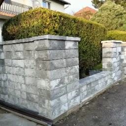 Nowoczesne ogrodzenie betonowe z szarej kostki modułowej, z widocznym systemem odprowadzania wody deszczowej i starannie przyciętymi tujami w tle.