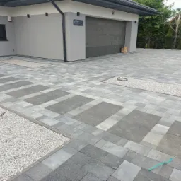 Ułożona kostka brukowa w dwóch odcieniach szarości, tworząca wzór prostokątów, z wysypanymi kamykami wzdłuż krawędzi i w specjalnie wydzielonych miejscach na podjeździe przed garażem.