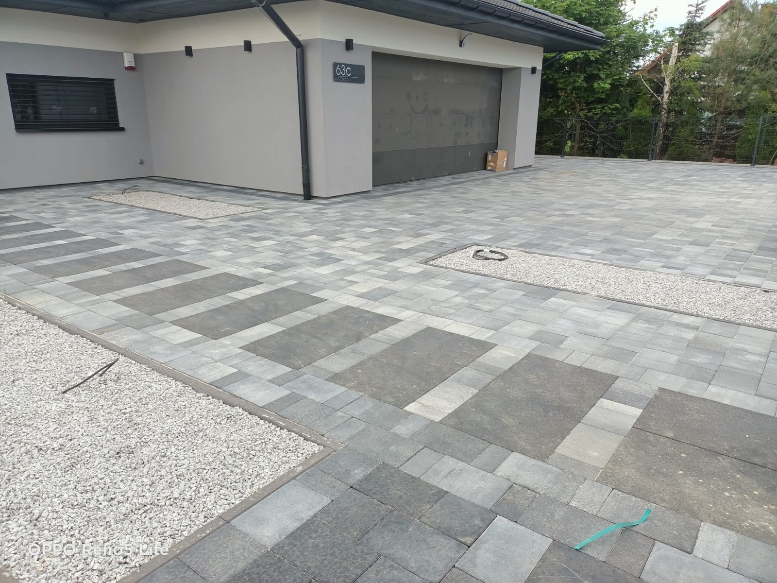 Ułożona kostka brukowa w dwóch odcieniach szarości, tworząca wzór prostokątów, z wysypanymi kamykami wzdłuż krawędzi i w specjalnie wydzielonych miejscach na podjeździe przed garażem.