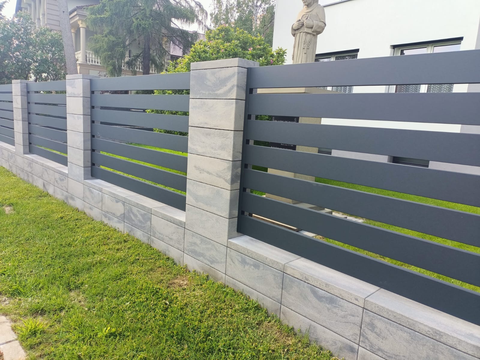 Nowoczesne ogrodzenie panelowe w kolorze grafitowym z podmurówką z szarych bloczków betonowych, widok z poziomu trawnika.