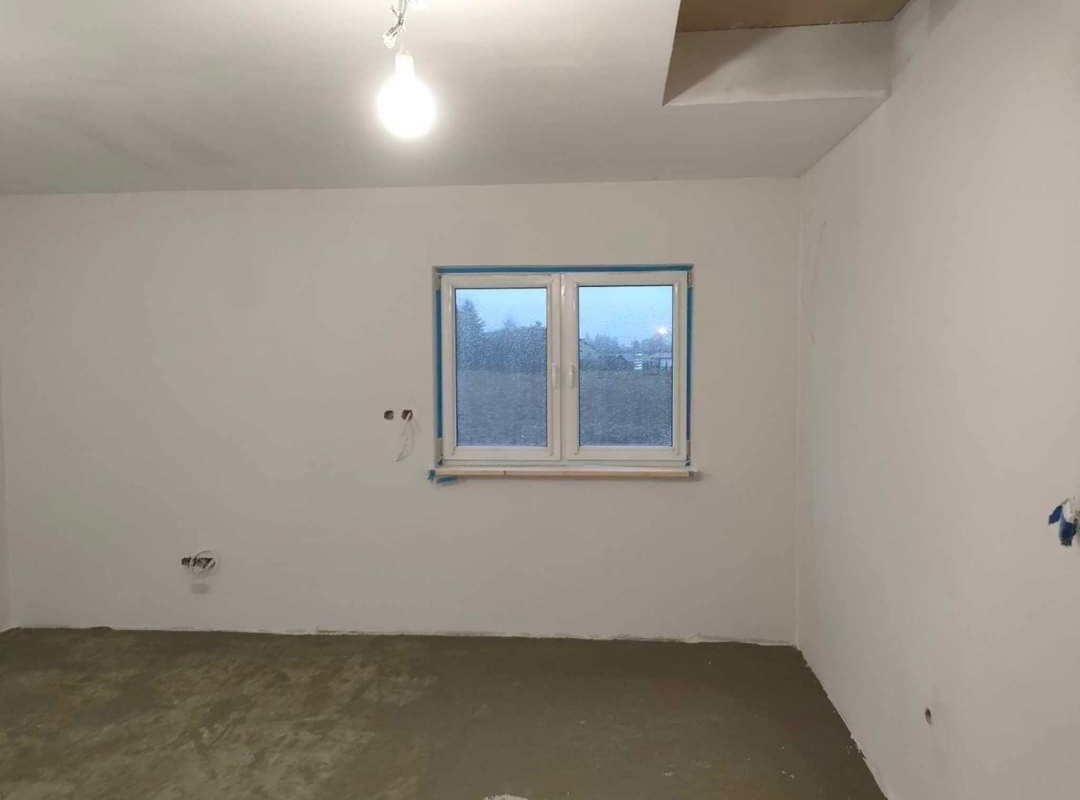 Pokój w trakcie prac wykończeniowych: świeżo położona wylewka na podłodze, ściany pokryte gładzią, okno zabezpieczone folią malarską, surowa żarówka zwisa z sufitu.