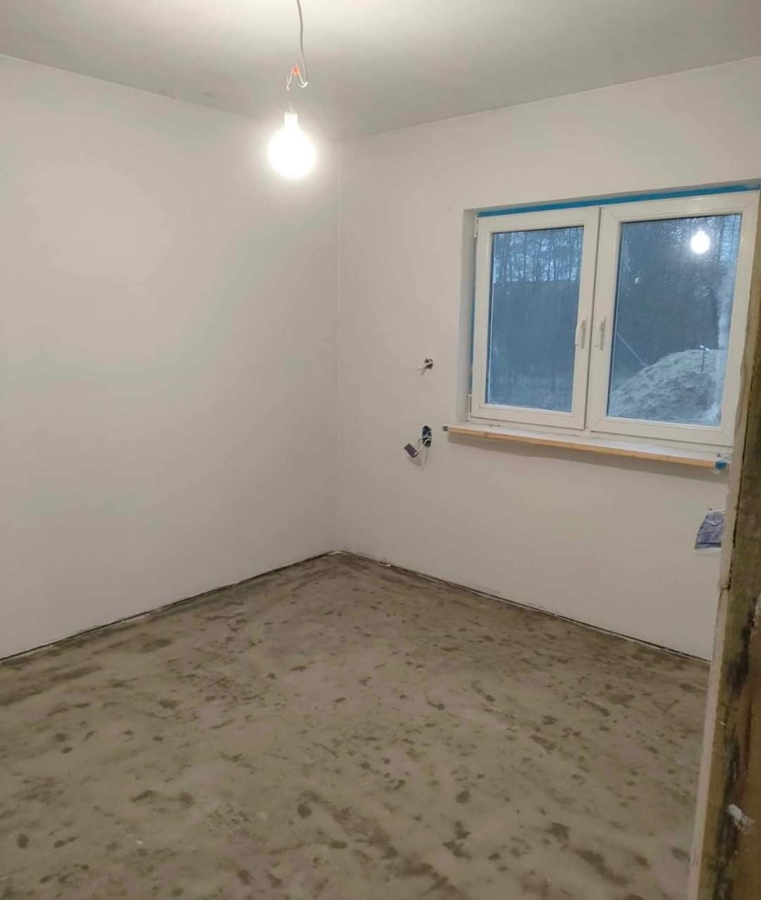 Pokój w stanie deweloperskim z surową podłogą, świeżo pomalowanymi na biało ścianami i oknem z widokiem na las. Na suficie zwisa pojedyncza żarówka.