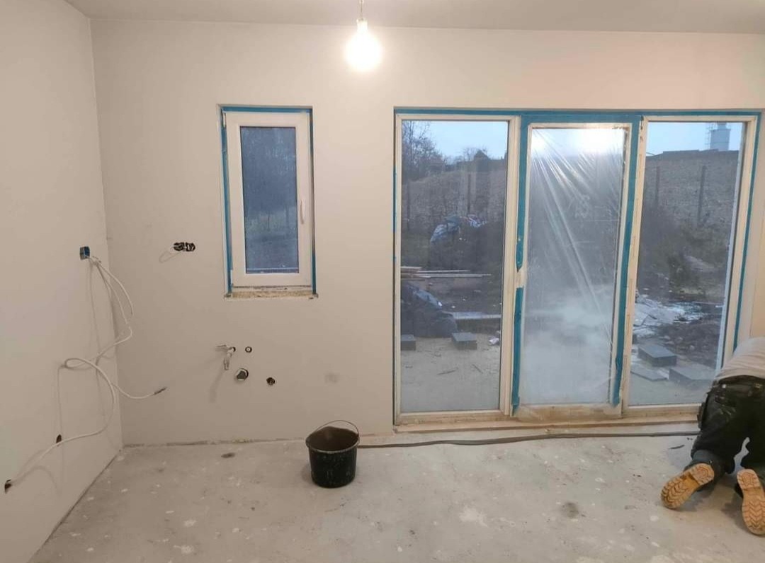 Wnętrze w trakcie prac wykończeniowych: ściany po nałożeniu gładzi, okna zabezpieczone folią, widoczne instalacje elektryczne, czarne wiadro na podłodze, fragment osoby w roboczym stroju.