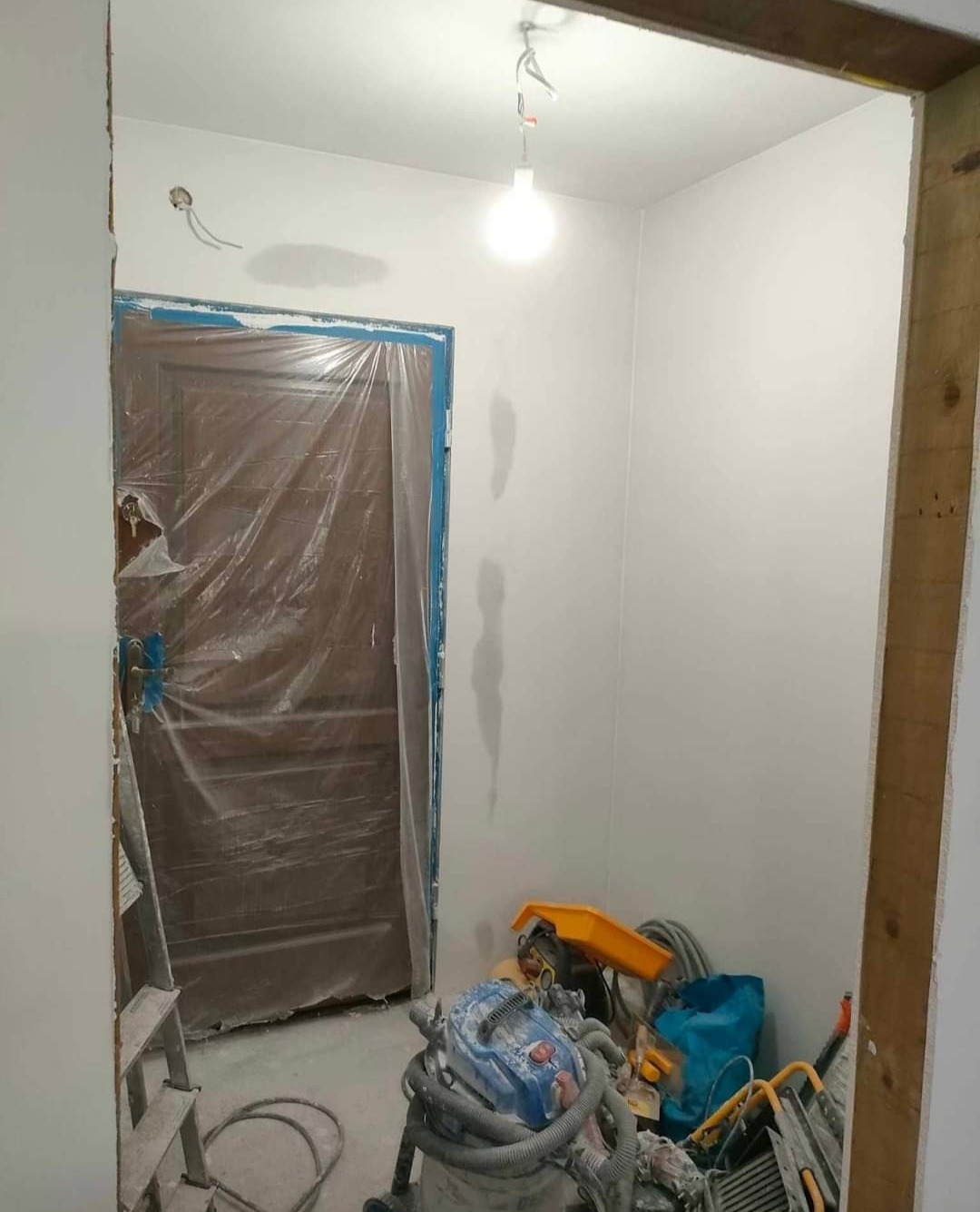 Pokój w trakcie odświeżania: zabezpieczone folią drzwi, świeżo pomalowane ściany z widocznymi niedoskonałościami, sprzęt malarski na podłodze, drabina oparta o ścianę.