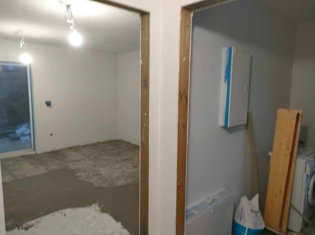 Przygotowanie do gładzi w Zasutowie: Widok przez futrynę na pokój z częściowo wylanym podkładem podłogowym i surowymi ścianami, widoczne instalacje elektryczne i grzewcze oraz pralka w tle.