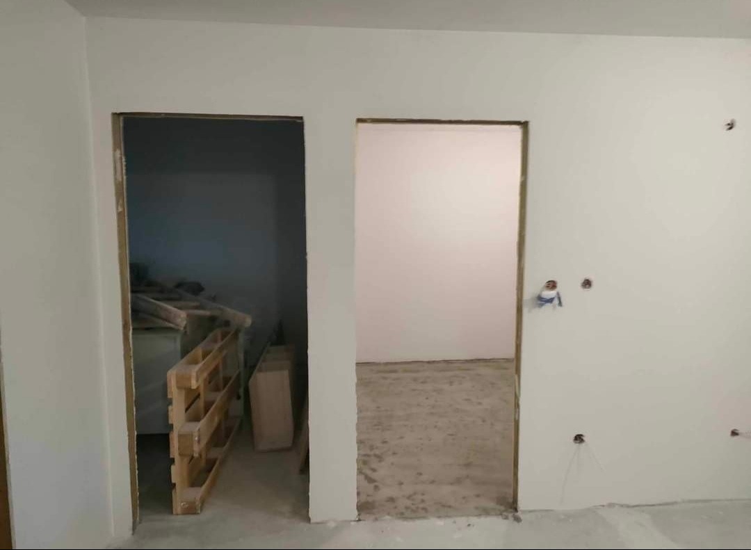 Wnętrze w trakcie prac wykończeniowych: surowe ściany po gładziowaniu, przygotowane otwory drzwiowe, widoczne palety w pomieszczeniu obok.