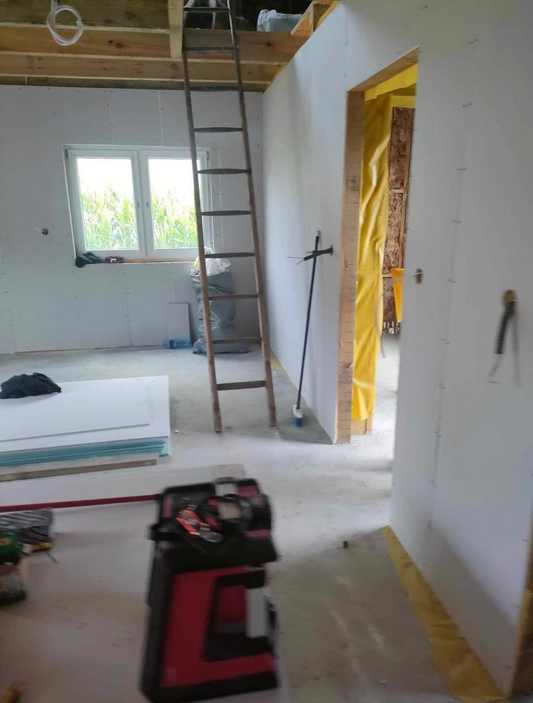 Wnętrze w trakcie prac wykończeniowych: ściany z płyt kartonowo-gipsowych, drewniana drabina oparta o ścianę, okno z widokiem na zewnątrz, narzędzia i materiały budowlane porozrzucane na podłodze...