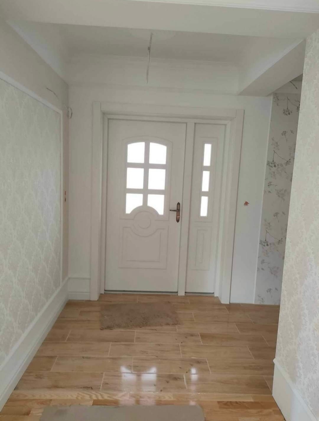 Białe drzwi wewnętrzne z szybkami, jasne płytki podłogowe imitujące drewno, wzorzysta tapeta na ścianach w przedpokoju po remoncie.