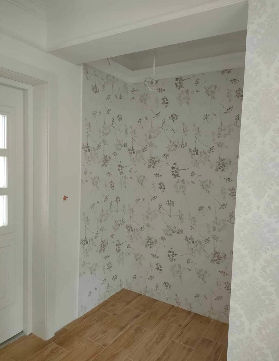 Wnętrze korytarza po remoncie: ściany pokryte tapetą w drobne kwiaty, drewnopodobna podłoga, białe futryny drzwi i fragment dekoracyjnej tapety z motywem roślinnym na ścianie po prawej.