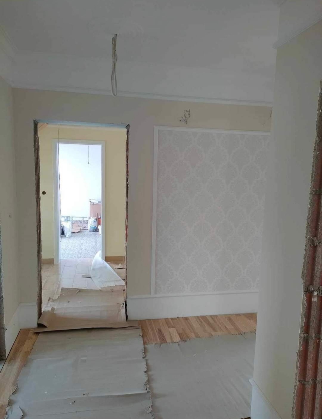 Pokój w trakcie prac wykończeniowych: położona podłoga drewniana zabezpieczona kartonem, ściany częściowo pomalowane, dekoracyjna tapeta w ramie, wystające przewody z sufitu, widoczne ślady po...