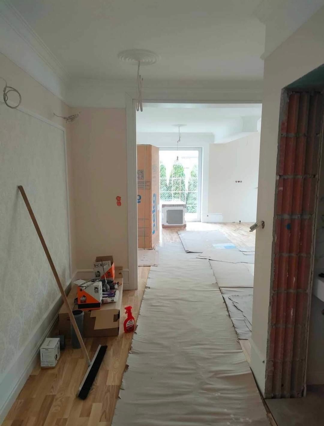 Korytarz w trakcie remontu: zabezpieczona podłoga, narzędzia i materiały budowlane, widok na odnowione pomieszczenie z oknem na ogród.