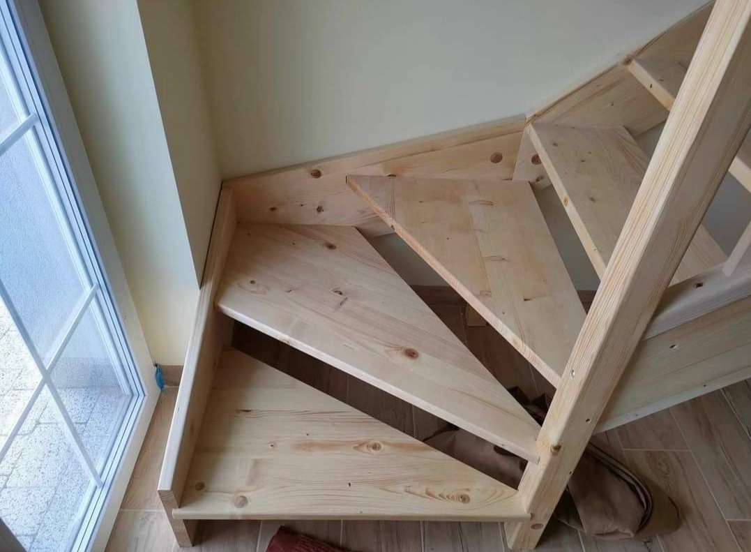 Surowe, drewniane schody wewnętrzne z jasnego drewna, widoczne z dołu, z oknem w tle, przed wykończeniem.