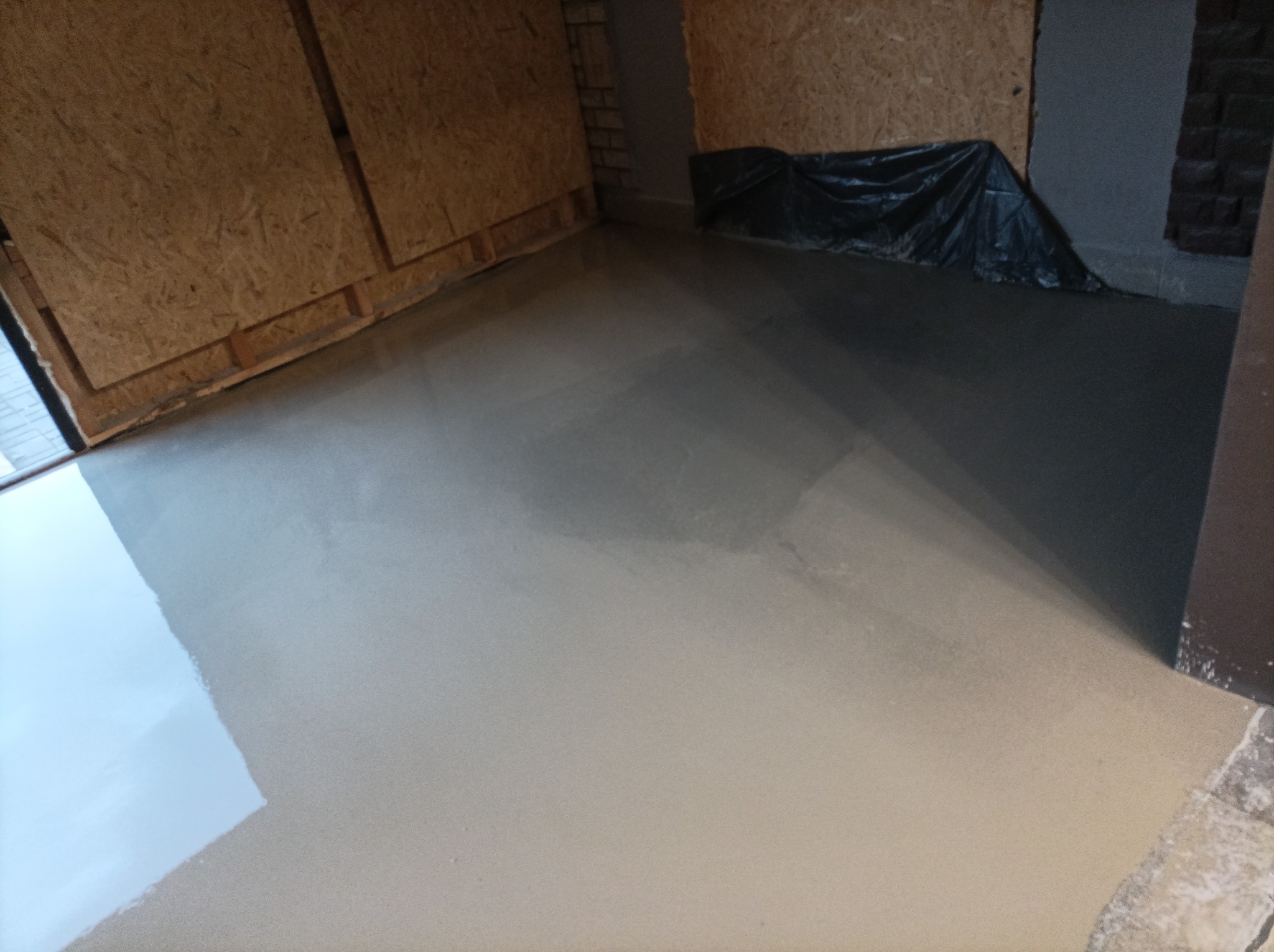 Świeżo wylana, gładka posadzka w pomieszczeniu z płytami OSB na ścianach i zabezpieczoną folią ścianą z cegły.