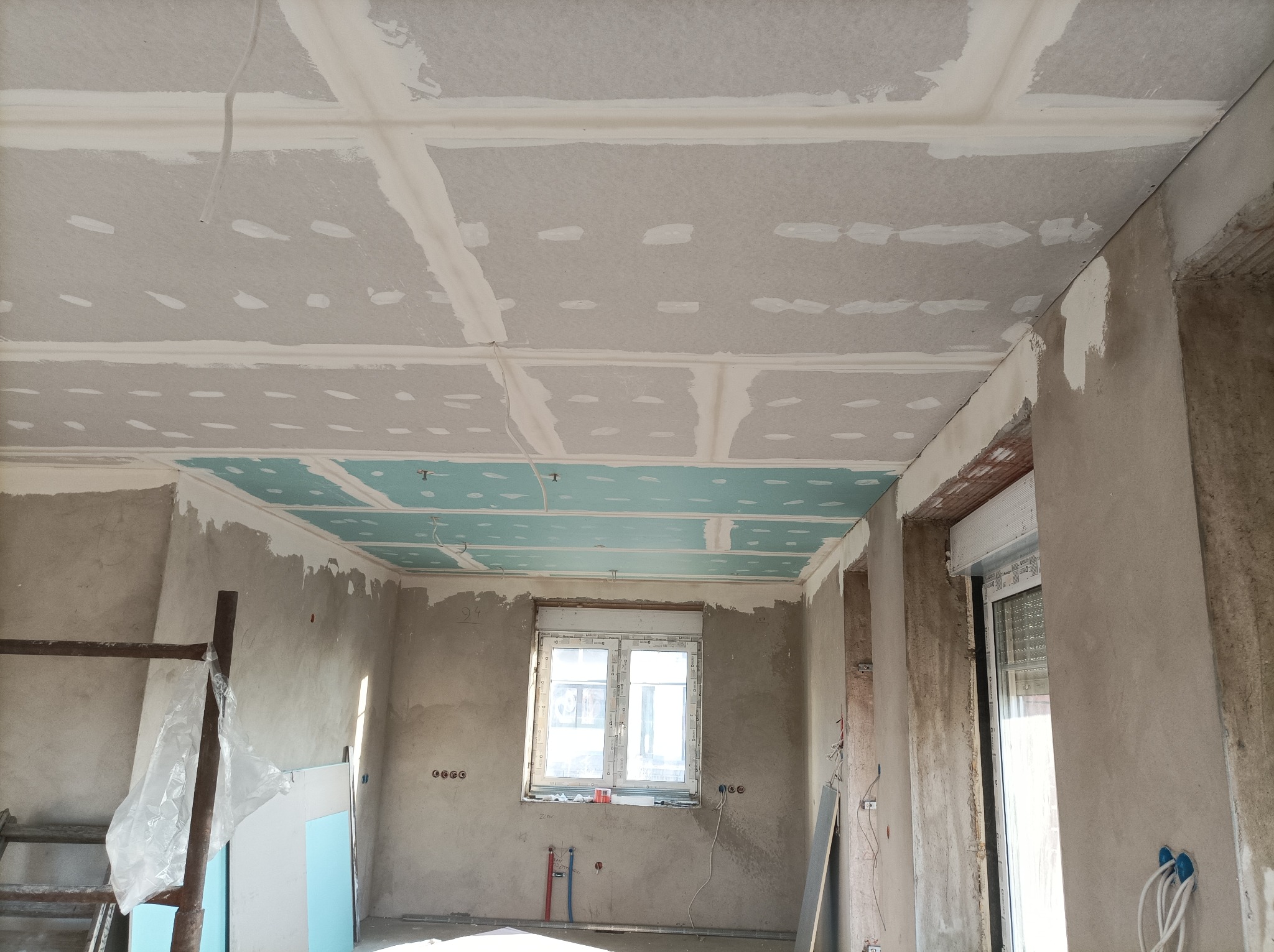 Sufit z płyt kartonowo-gipsowych, częściowo zagruntowany na niebiesko, z widocznymi łączeniami wypełnionymi szpachlą, na ścianach widoczne ślady po pracach remontowych, okno z białymi ramami.