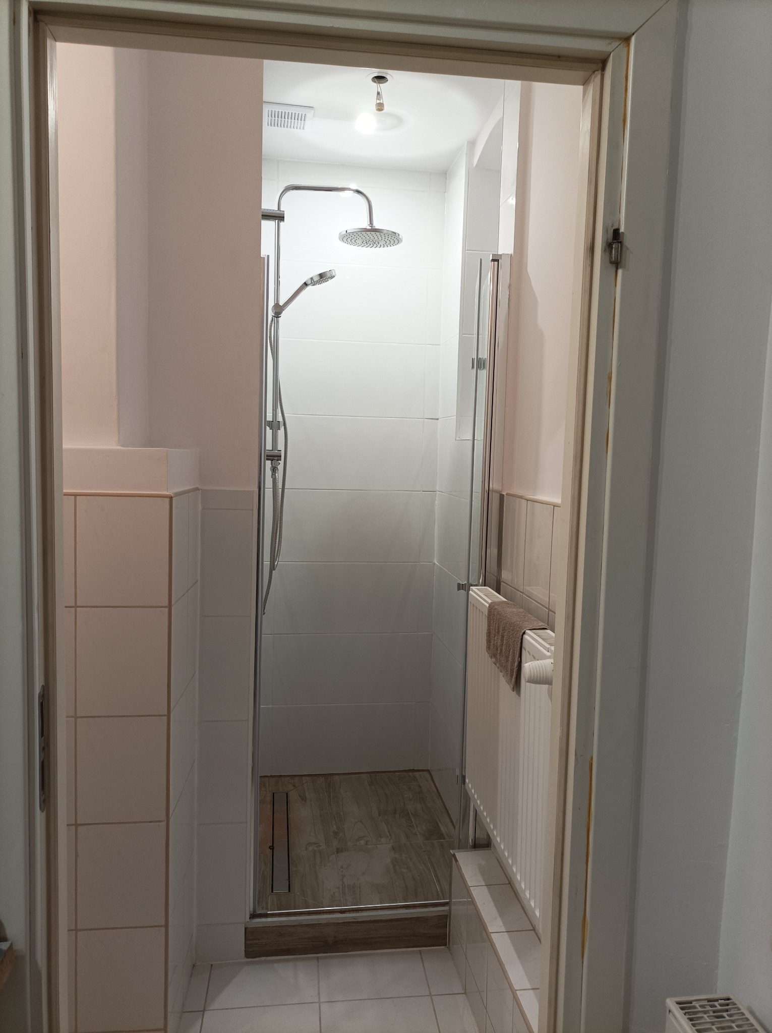 Wyremontowana łazienka z kabiną prysznicową typu walk-in, wyłożona białymi płytkami, z drewnopodobną podłogą w kabinie i odpływem liniowym, widok zza uchylonych drzwi.