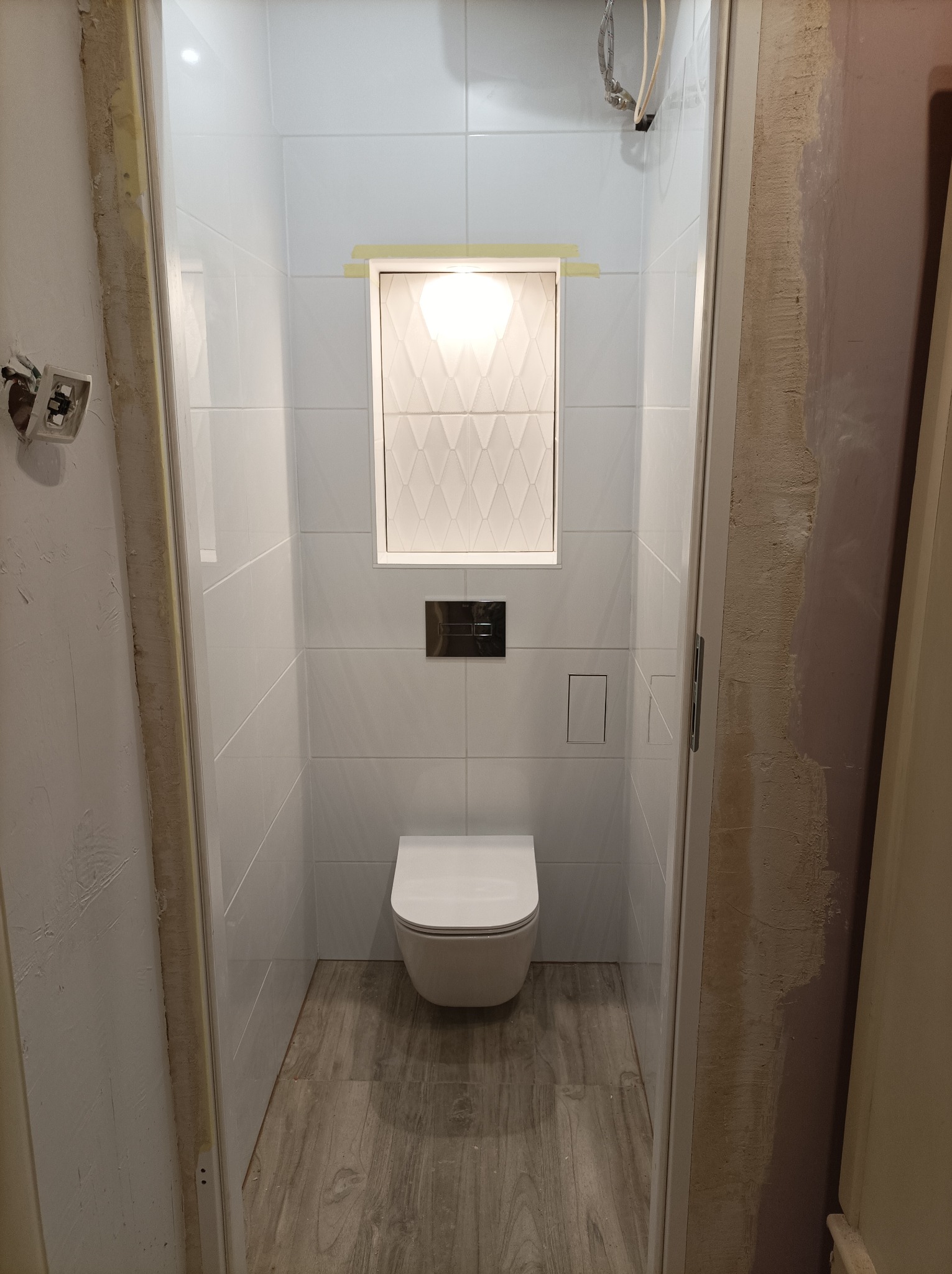 Wnętrze łazienki w trakcie remontu z białymi płytkami na ścianach, wiszącą toaletą i panelami podłogowymi imitującymi drewno, widoczne niedokończone obrzeża drzwi.