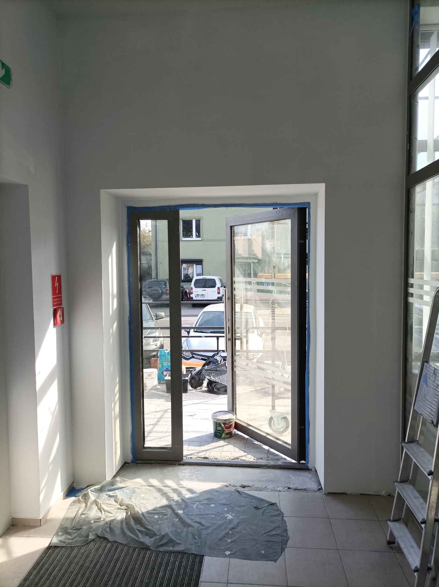 Wnętrze w trakcie odświeżania: świeżo pomalowane ściany na biało, zabezpieczone folią podłogi, widoczne otwarte drzwi na zewnątrz i drabina malarska.