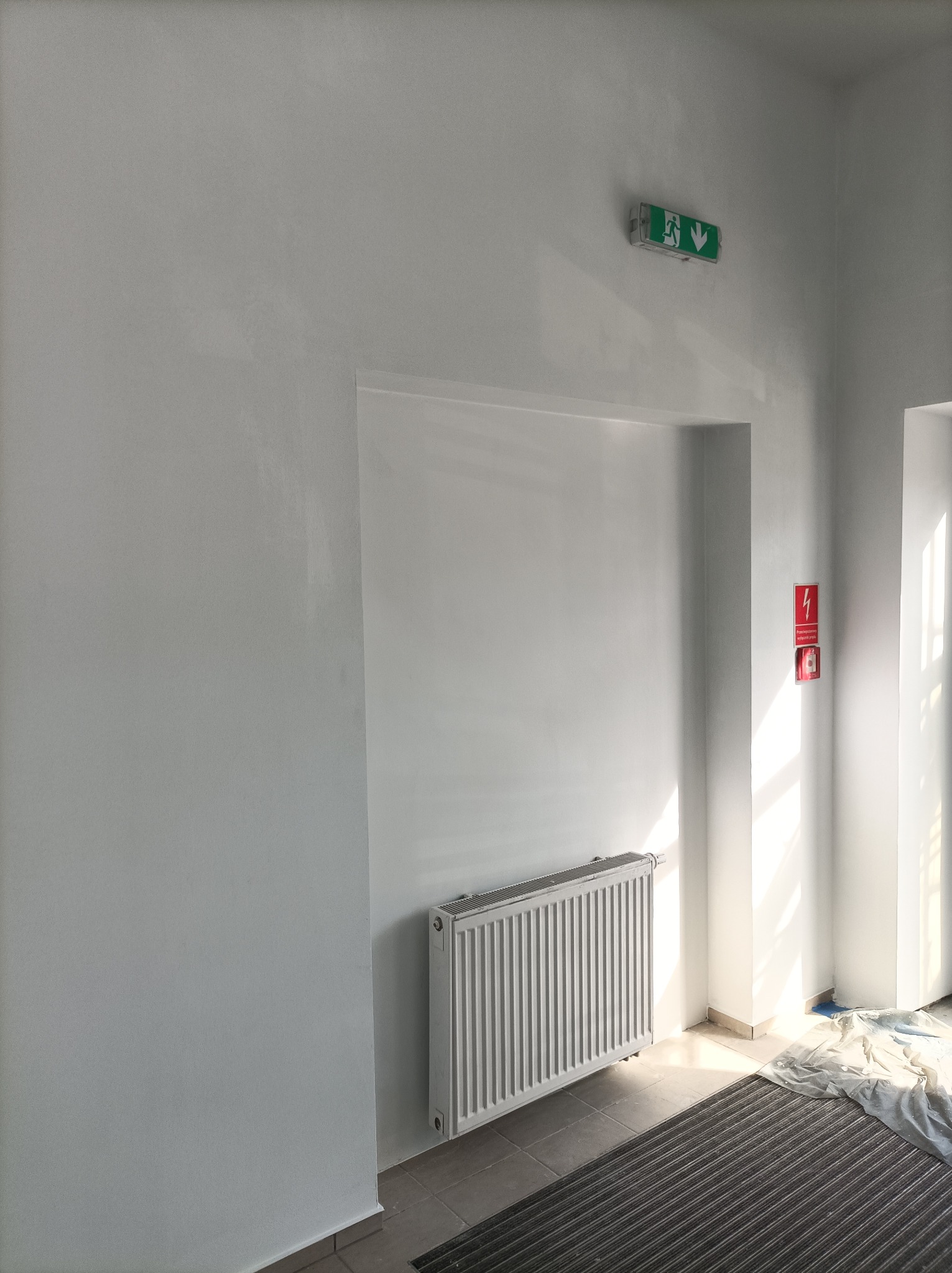 Świeżo pomalowane na biało ściany w korytarzu budynku, widoczny grzejnik, czujnik dymu i znak ewakuacyjny, podłoga częściowo zabezpieczona folią malarską.