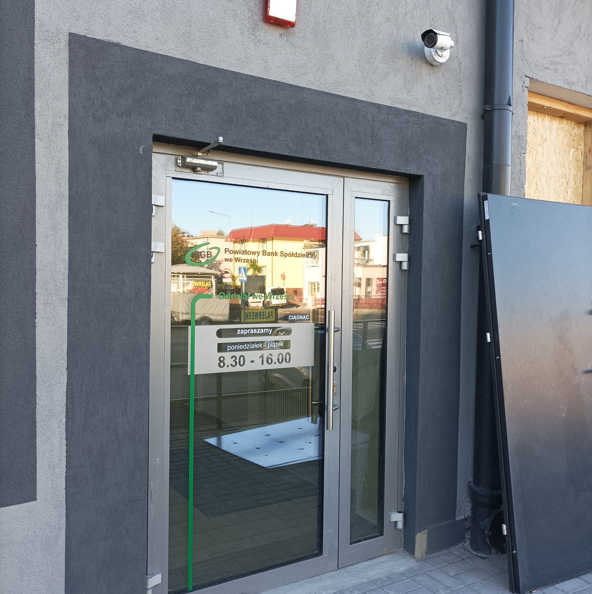Wejście do oddziału banku z szarą elewacją, widoczna kamera monitoringu oraz drzwi z informacją o godzinach otwarcia.