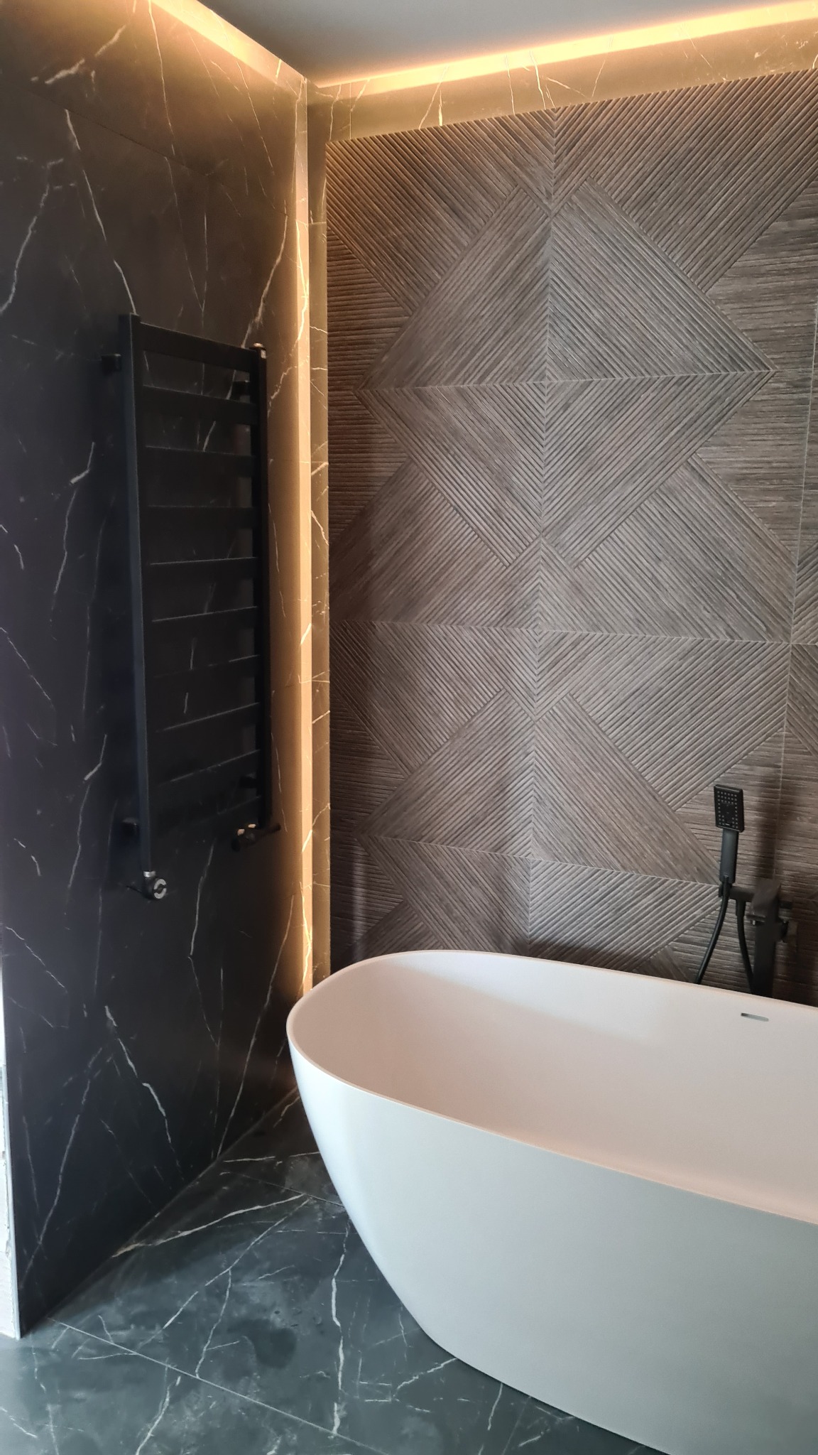 Nowoczesna łazienka z wolnostojącą białą wanną, czarnym grzejnikiem drabinkowym na ścianie z imitacją czarnego marmuru oraz dekoracyjną ścianą z geometrycznym wzorem, podświetlona listwą LED.