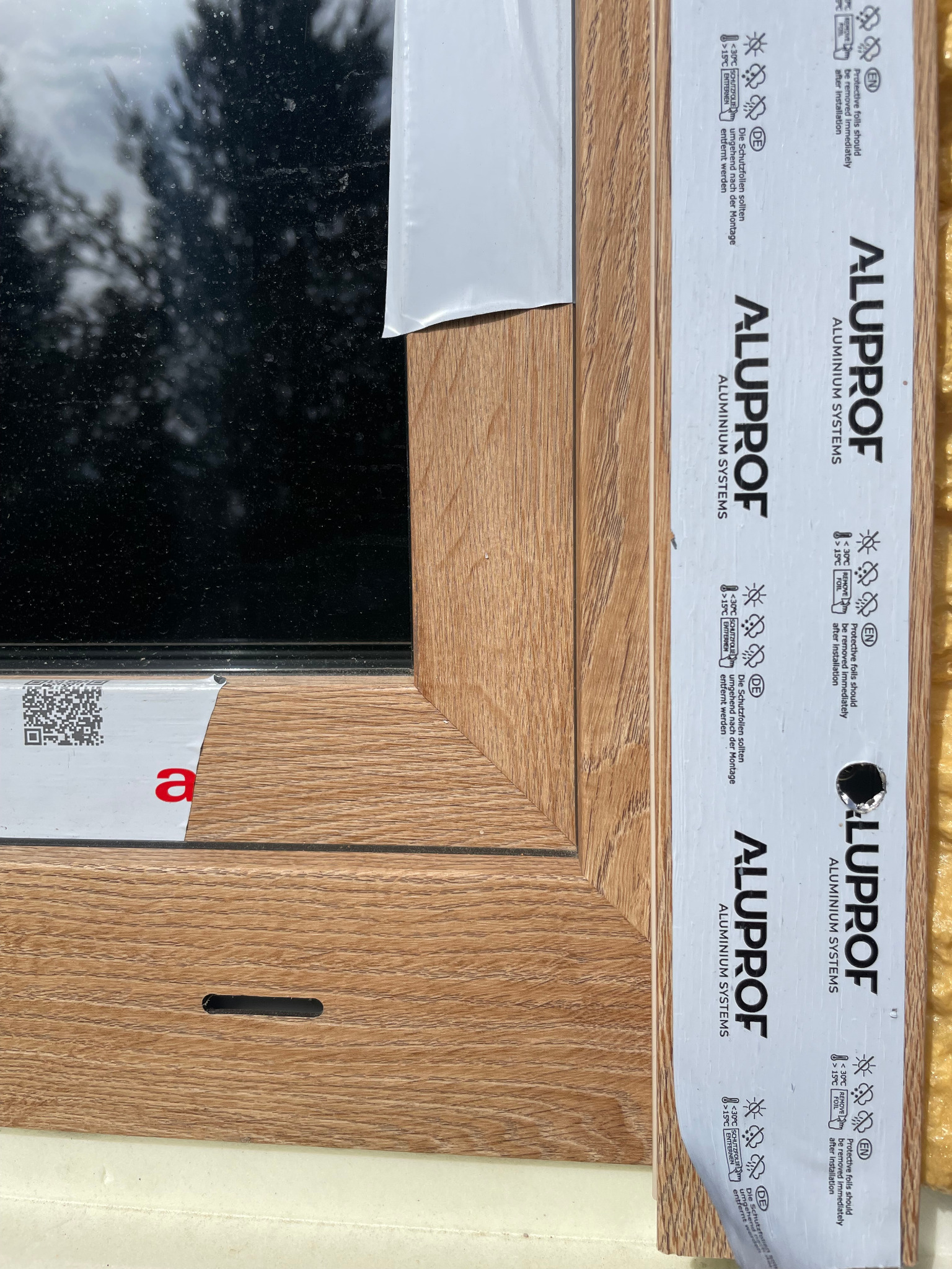 Fragment okna z drewnopodobną ramą, zabezpieczoną folią ochronną Alupof. Widoczny profil okienny i szyba. Detal montażu okna z bliska.