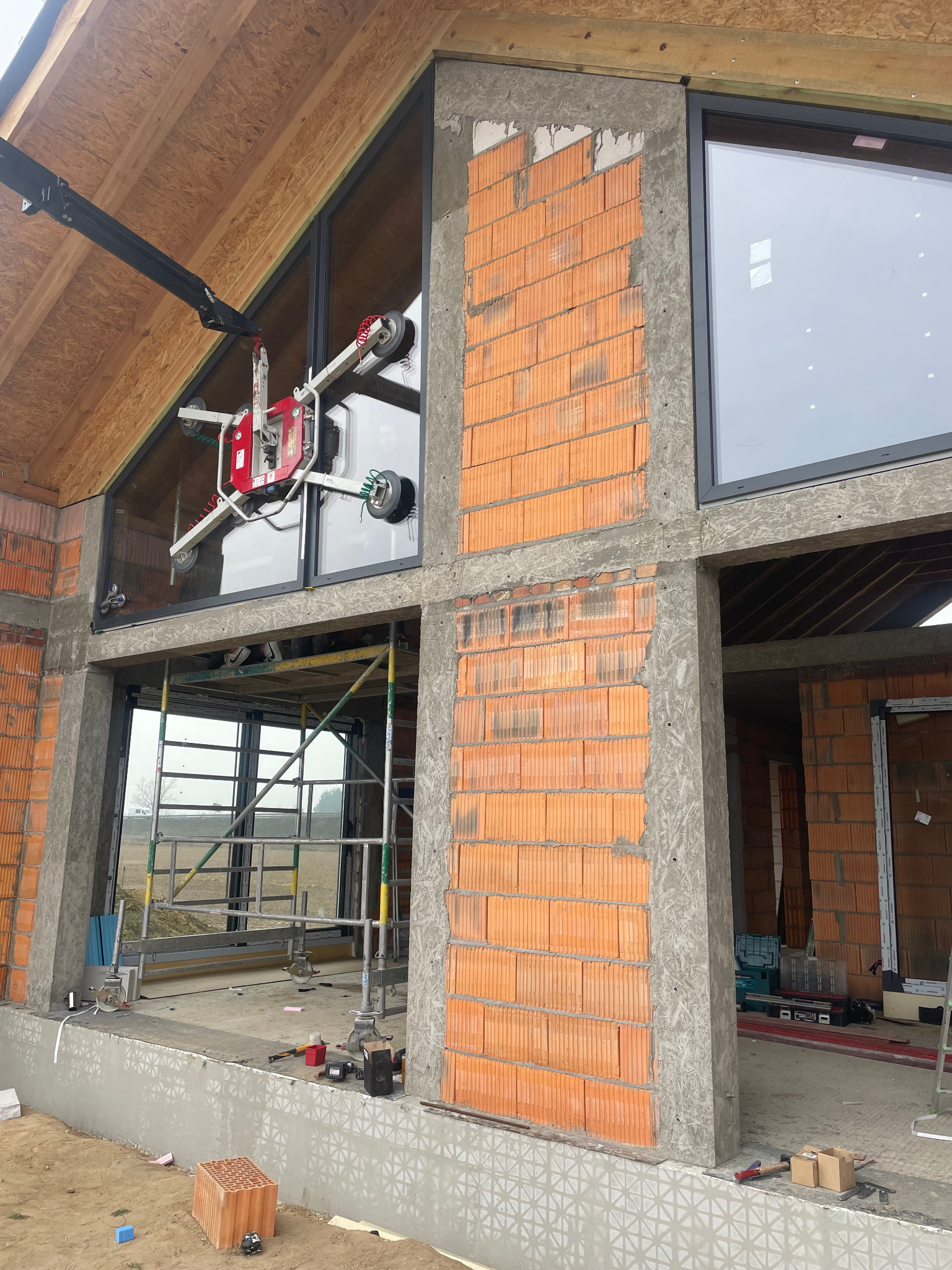 Montaż okna z użyciem specjalistycznego podnośnika przyssawkowego na budowie domu z cegły. Widoczne rusztowania i narzędzia.