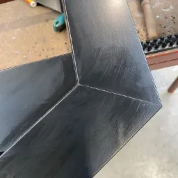 Szczegół narożnika czarnej ramy okiennej w warsztacie w Cikowicach, widoczne ślady obróbki i narzędzia na stole roboczym.