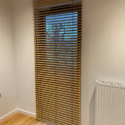 Drewniane żaluzje poziome zamontowane w oknie, widok na zewnątrz z drzewami, biała ściana i grzejnik w tle.