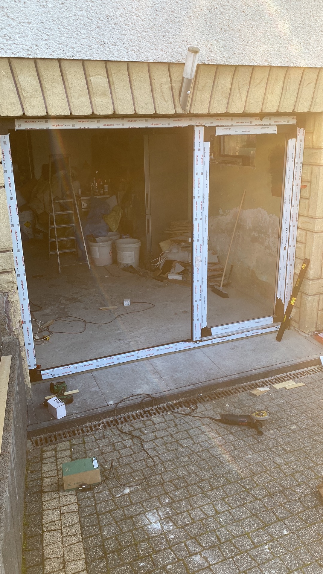 Montaż ramy okiennej z białego PCV w garażu, widoczne narzędzia pracy, poziomica i szlifierka kątowa na zewnątrz, wnętrze garażu w trakcie prac porządkowych.