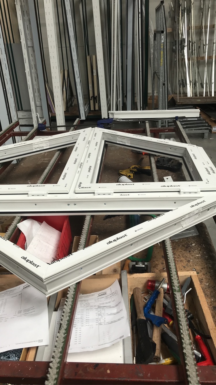 Białe ramy okienne Aluplast ułożone na metalowym stole warsztatowym w hali produkcyjnej, widoczne narzędzia i dokumentacja techniczna.