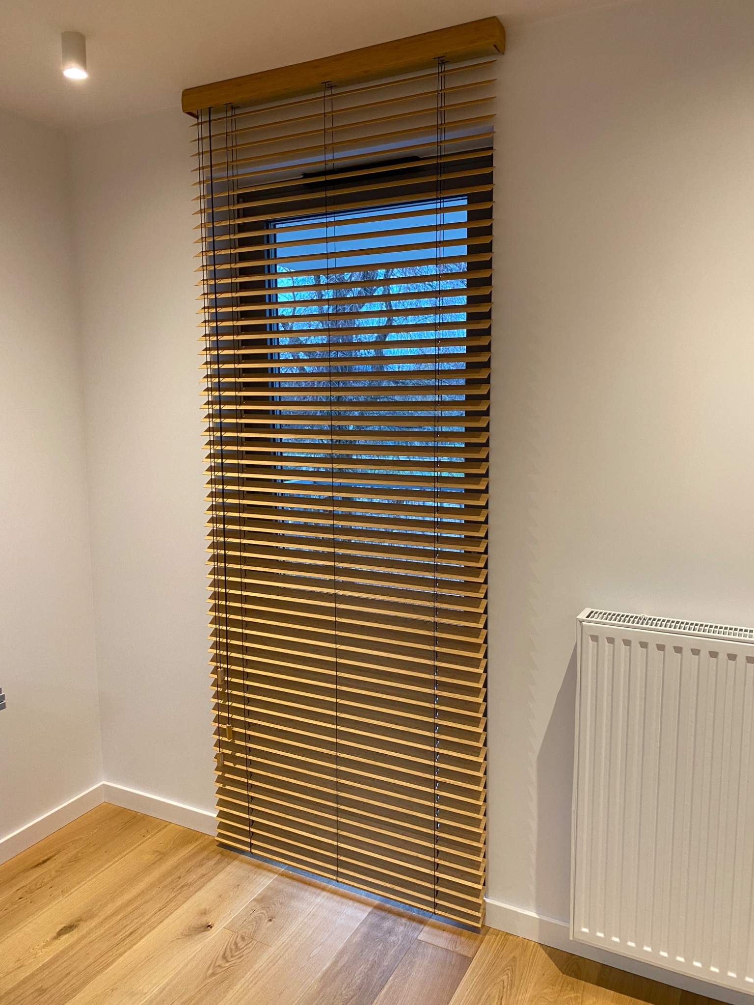 Drewniane żaluzje poziome zamontowane w oknie, widok na zewnątrz z drzewami, biała ściana i grzejnik w tle.