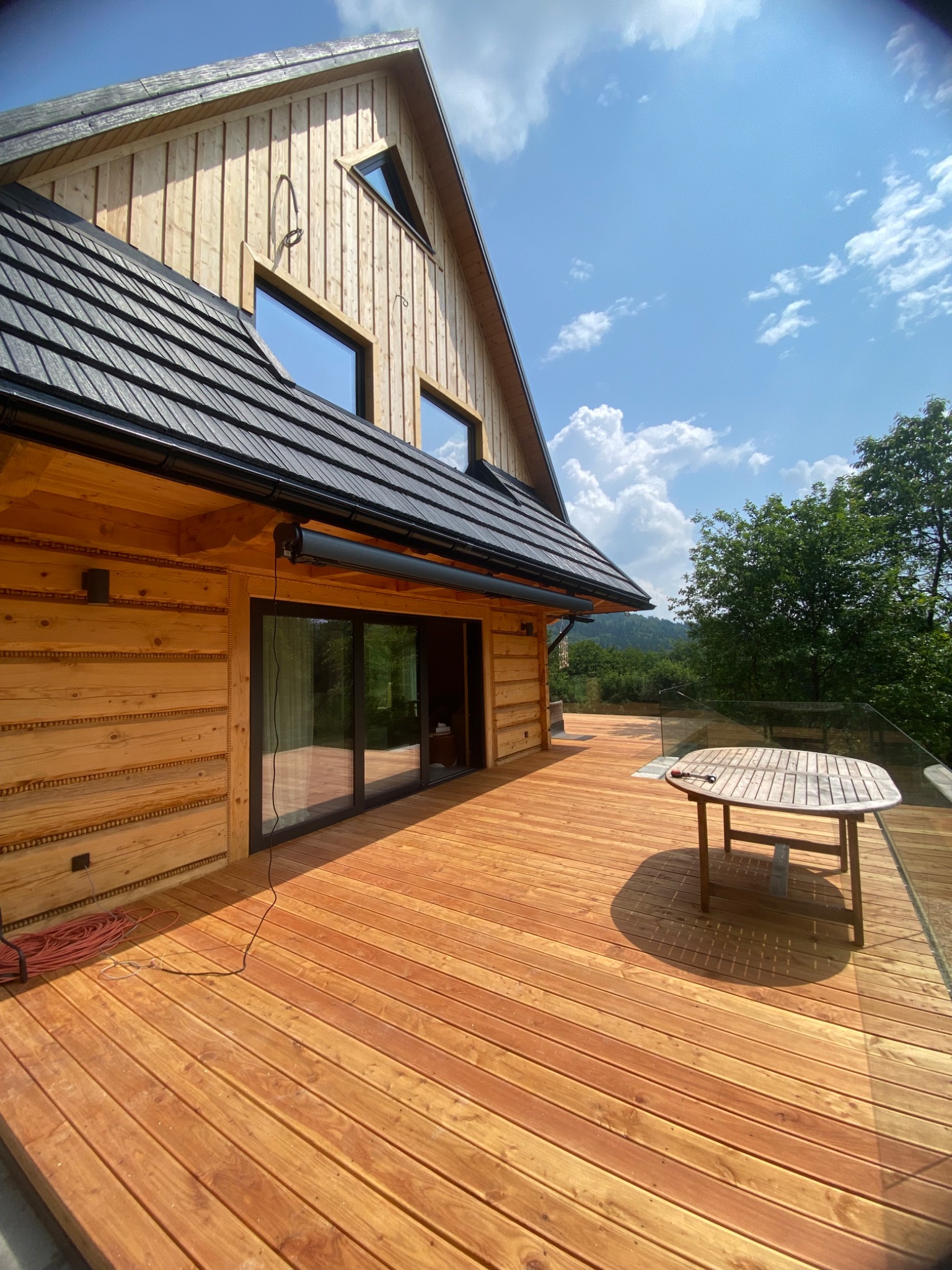 Drewniany dom z tarasem, widoczne okna, roleta zwinięta nad oknem tarasowym, stół na tarasie, błękitne niebo w tle.