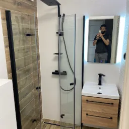 Nowoczesna łazienka z kabiną prysznicową typu walk-in, drewnopodobnymi płytkami na ścianie i podłodze, oraz szafką pod umywalką z drewnianym fornirem i czarnymi uchwytami. W lustrze odbija się...