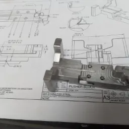 Metalowy blok 'PUSHER BLOCK' wykonany ze stali nierdzewnej 304, leżący na rysunku technicznym z wymiarami i oznaczeniami, prezentujący efekt obróbki CNC.