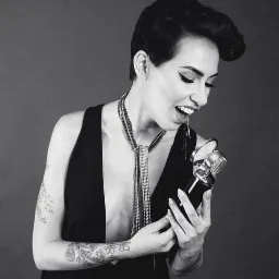 Czarno-białe zdjęcie kobiety z krótkimi włosami, w czarnej sukience z głębokim dekoltem, trzymającej mikrofon i śpiewającej, z widocznymi tatuażami na ramionach i długim, srebrnym naszyjnikiem...