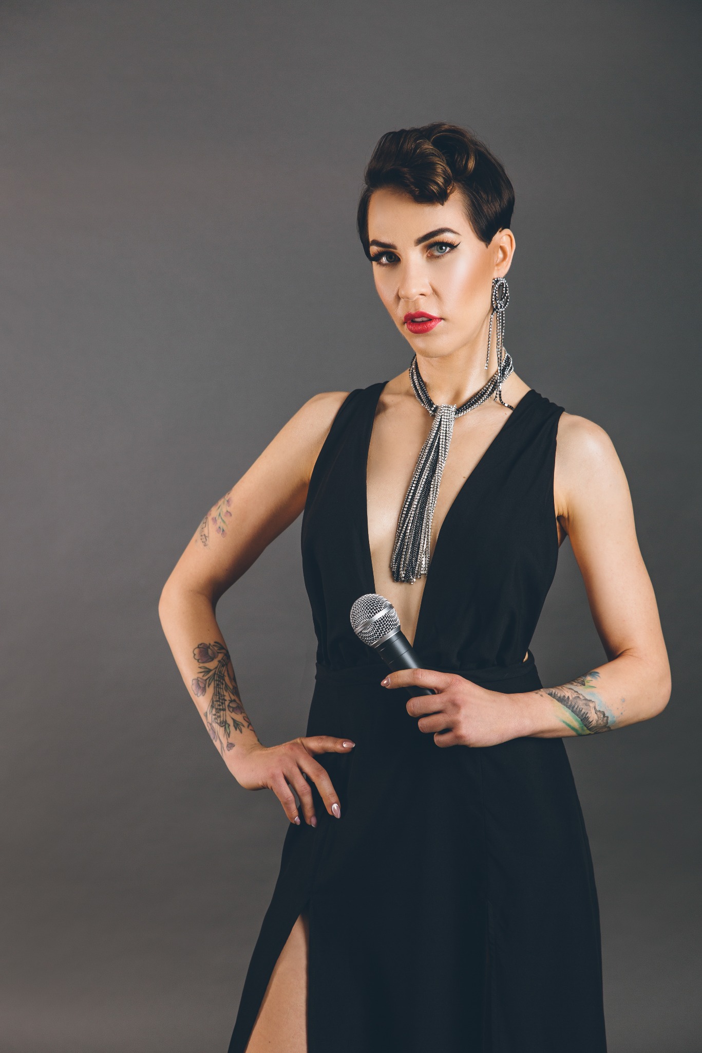 Kobieta z krótkimi włosami, w czarnej sukni z głębokim dekoltem i srebrnej biżuterii, trzyma mikrofon, pozuje na szarym tle, eksponując tatuaże na ramionach.