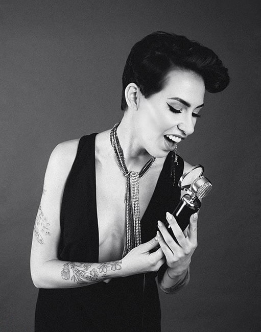 Czarno-białe zdjęcie kobiety z krótkimi włosami, w czarnej sukience z głębokim dekoltem, trzymającej mikrofon i śpiewającej, z widocznymi tatuażami na ramionach i długim, srebrnym naszyjnikiem...