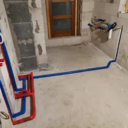 Surowe wnętrze z instalacją wodno-kanalizacyjną w trakcie montażu: czerwone i niebieskie rury prowadzone po ścianach z bloczków, widoczne zawory i filtr wody.