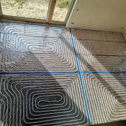 Instalacja ogrzewania podłogowego z rur ułożonych w pętle na folii aluminiowej, widoczne okno dachowe i fragment ściany.