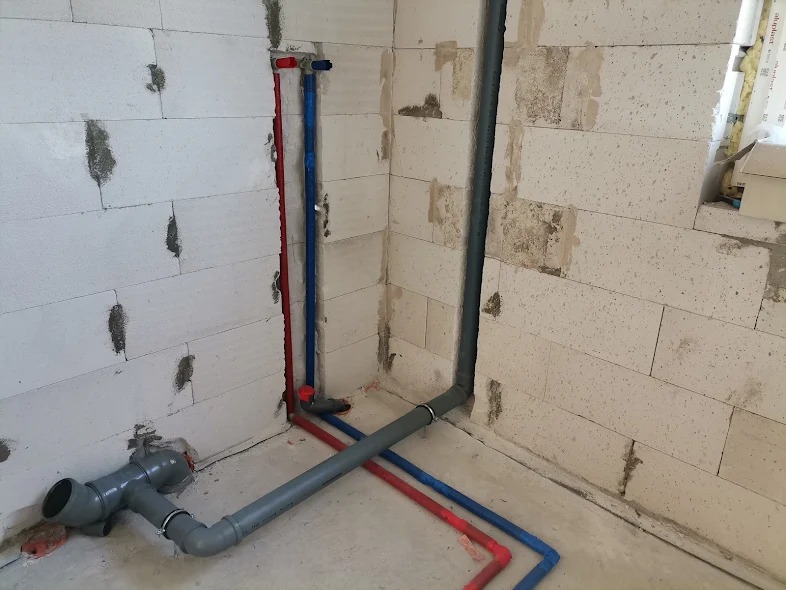 Surowe wnętrze z instalacją wodno-kanalizacyjną w trakcie montażu: szare rury kanalizacyjne, czerwone i niebieskie rury doprowadzające wodę, widoczne połączenia i mocowania na tle białych bloczków.