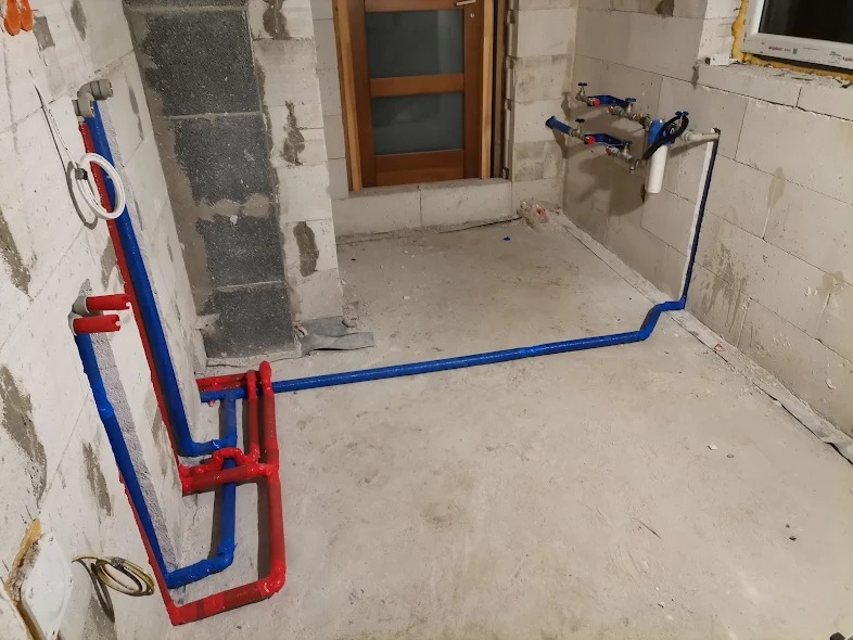 Surowe wnętrze z instalacją wodno-kanalizacyjną w trakcie montażu: czerwone i niebieskie rury prowadzone po ścianach z bloczków, widoczne zawory i filtr wody.