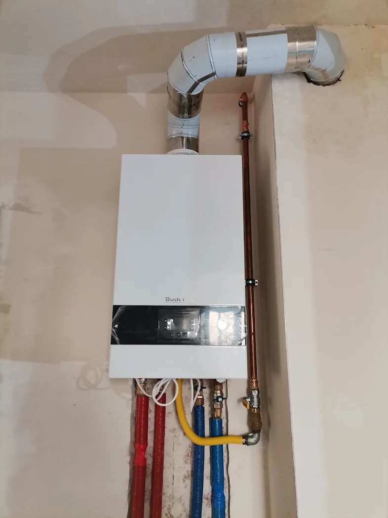 Biały piec gazowy marki Buderus zamontowany na ścianie, z widocznymi rurami doprowadzającymi i odprowadzającymi, zabezpieczonymi izolacją.